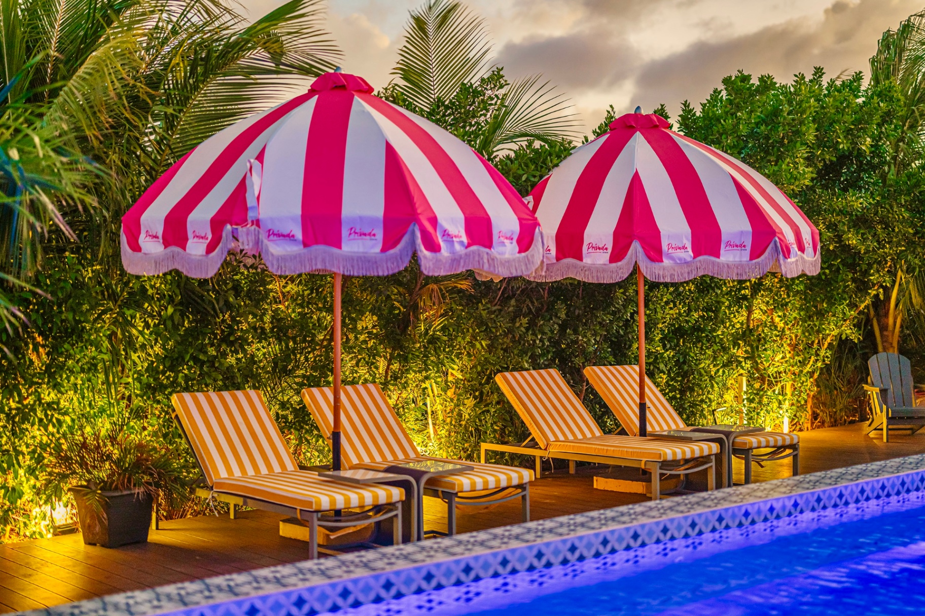 Two pink-and-white Privada-branded fringed parasols over orange loungers at dusk