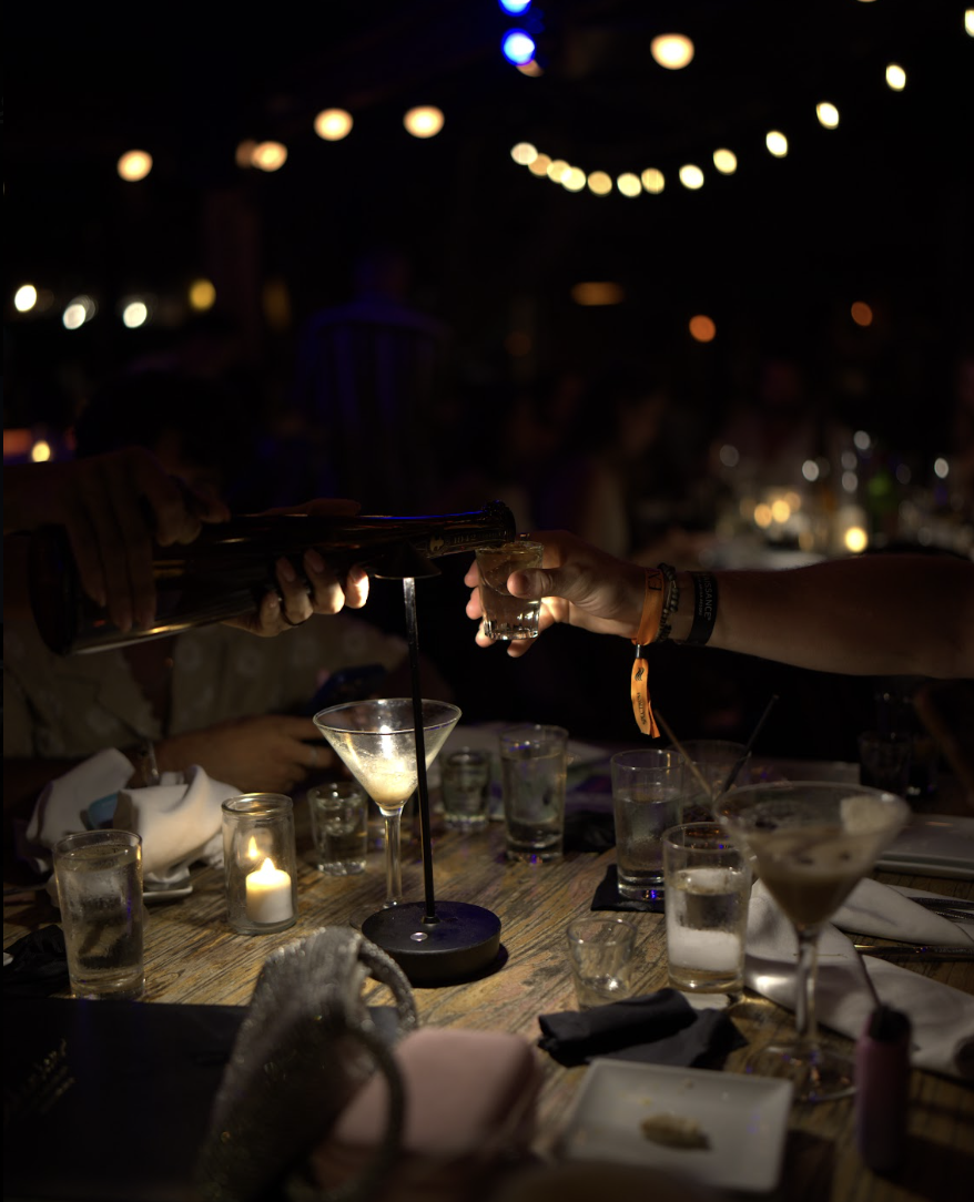 A bottle and a shot glass meet across a candlelit dinner table